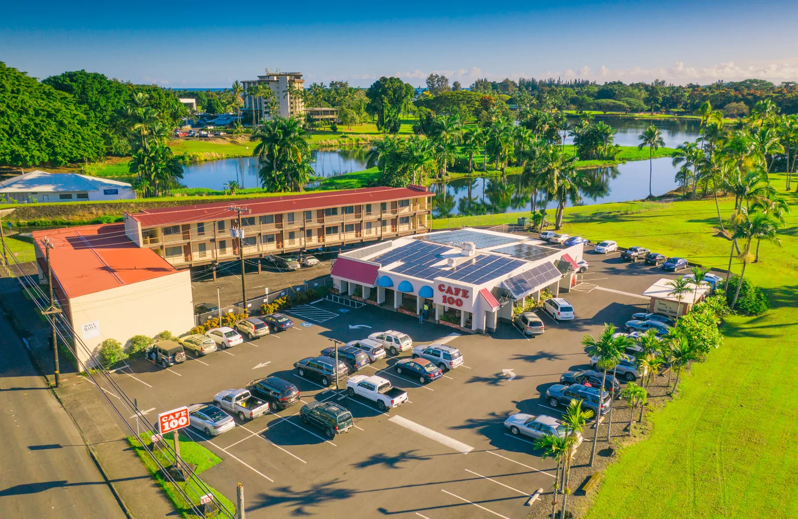 Aerial view of the Cafe 100 drive-in on Kilauea Avenue in Hilo, with its red Cafe 100 signage, blue awnings and full parking lot beside the Wailoa pond and palms.