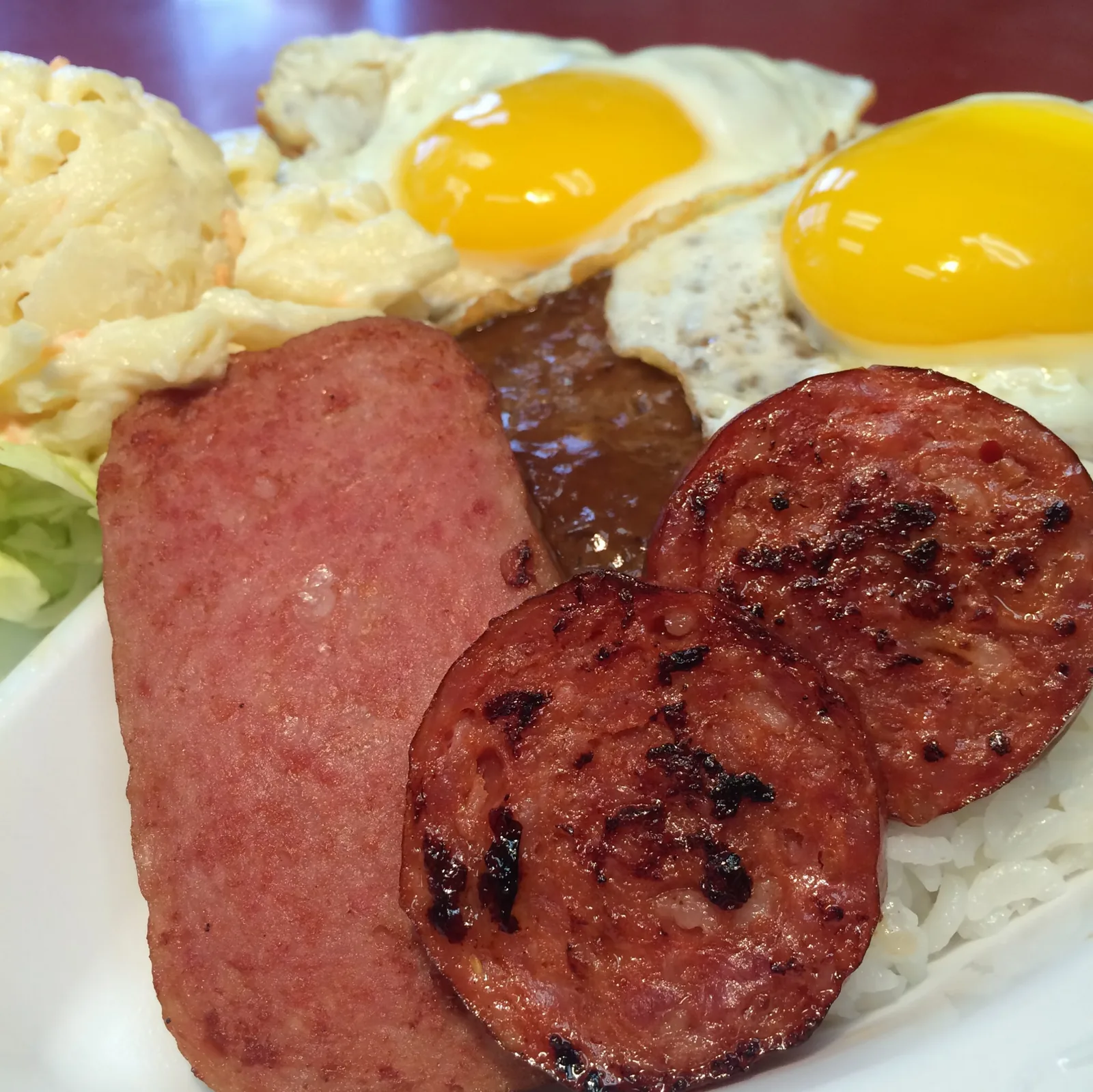 A loaded Cafe 100 plate: fried Spam, grilled Portuguese sausage, a hamburger patty under brown gravy, two sunny-side-up eggs, macaroni salad and rice.