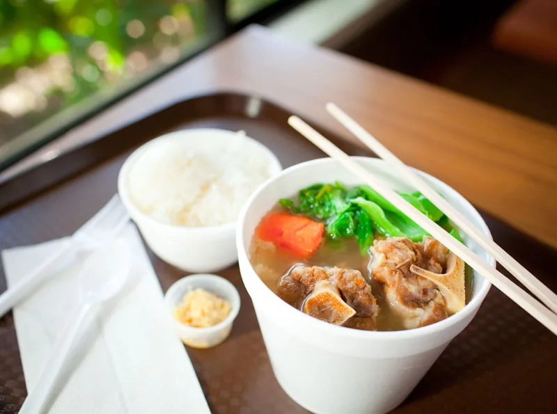 A cup of Cafe 100 oxtail soup with oxtail, carrot and choi sum in broth, served with a side of rice.